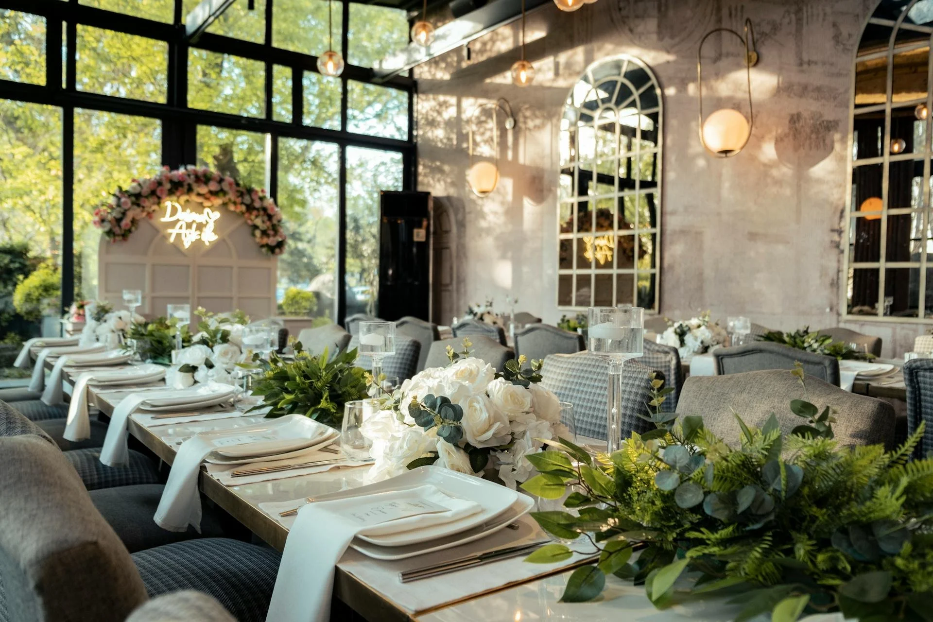 Decorated wedding reception table with white flowers, greenery, candles in glass holders, and white place settings in a bright indoor venue with large windows and modern lighting.