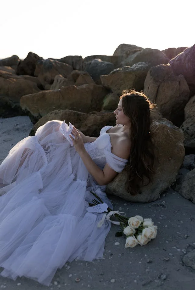A woman in a white wedding dress sitting on the rocky beach, holding a piece of paper, with white roses placed next to her, during sunset.