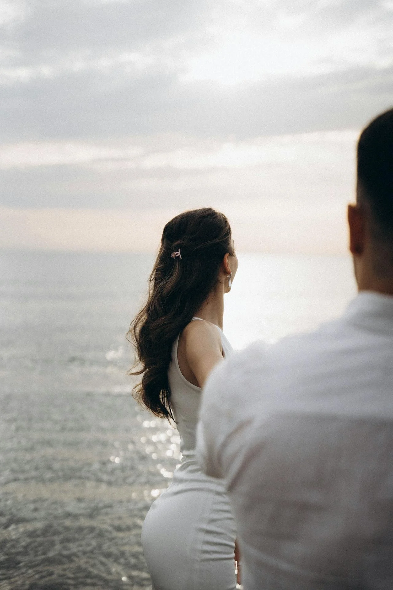 A woman in a white dress with long dark hair standing near the ocean, facing away, with a man partially visible beside her, during sunset or overcast weather.