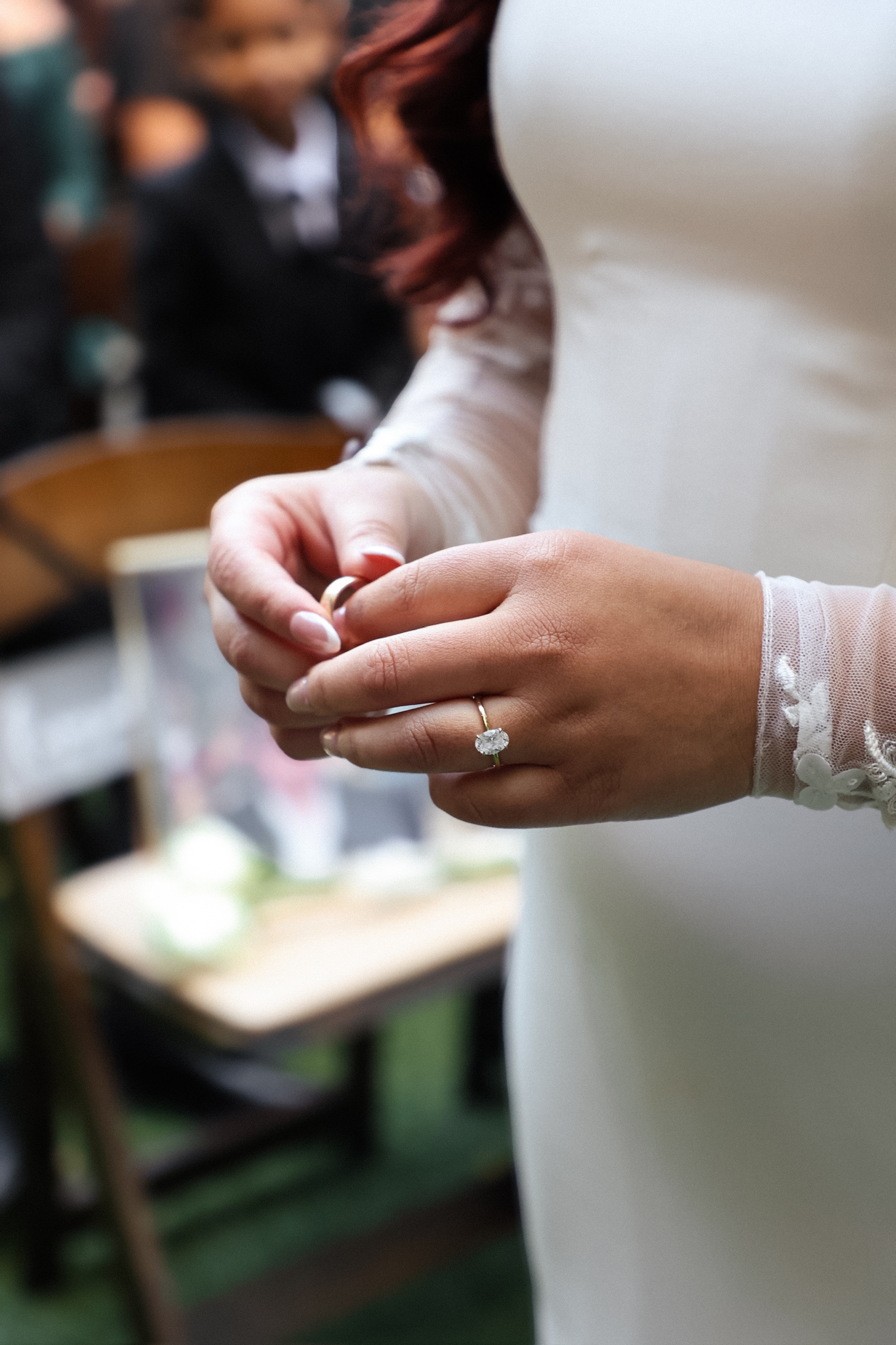 A woman wearing a white dress with lace sleeves is holding a wedding ring, preparing to place a ring on her finger during a wedding ceremony.