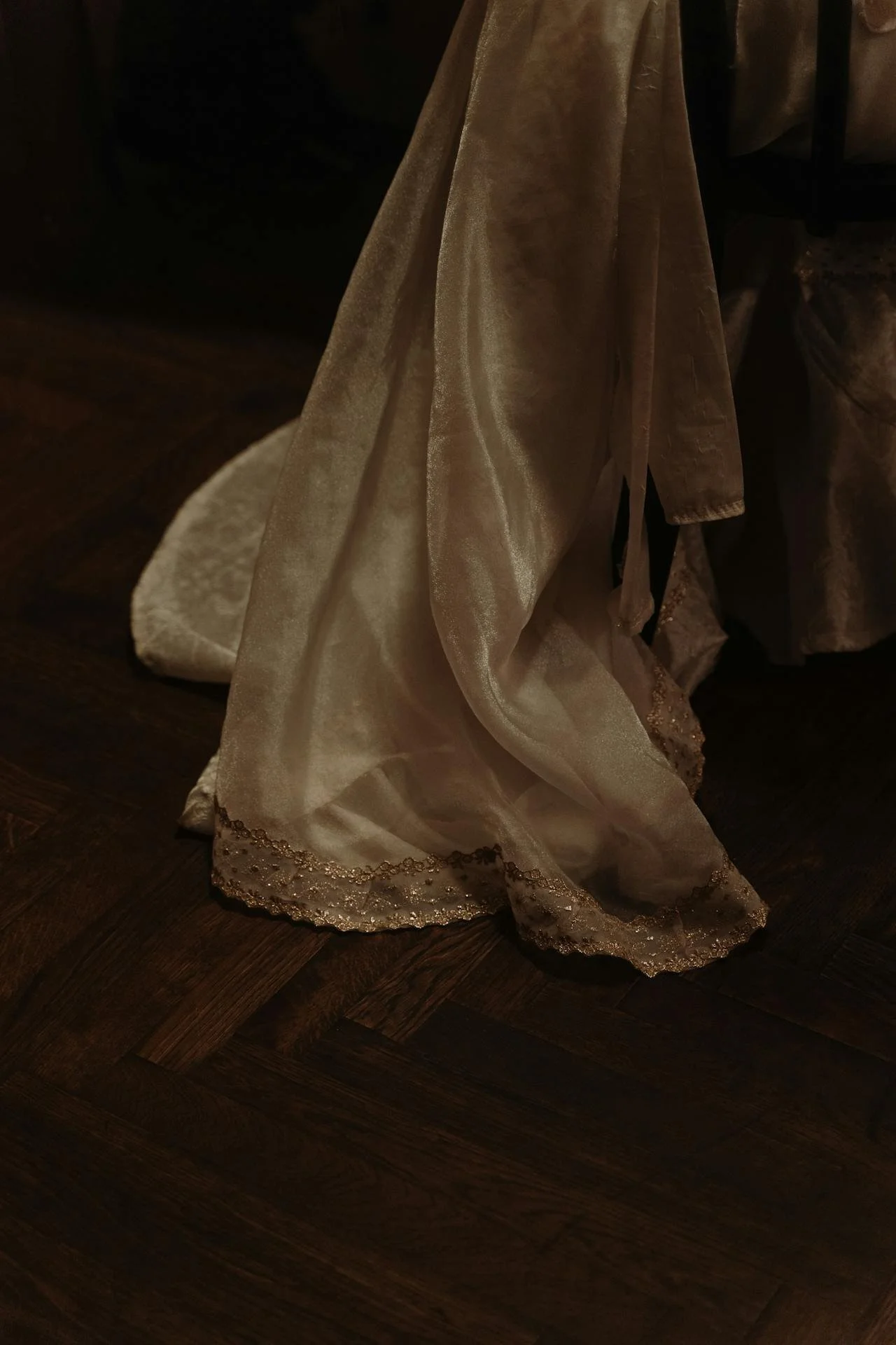 Close-up of a flowing, beige satin fabric with lace trim at the bottom, draped against a dark background and wooden floor.