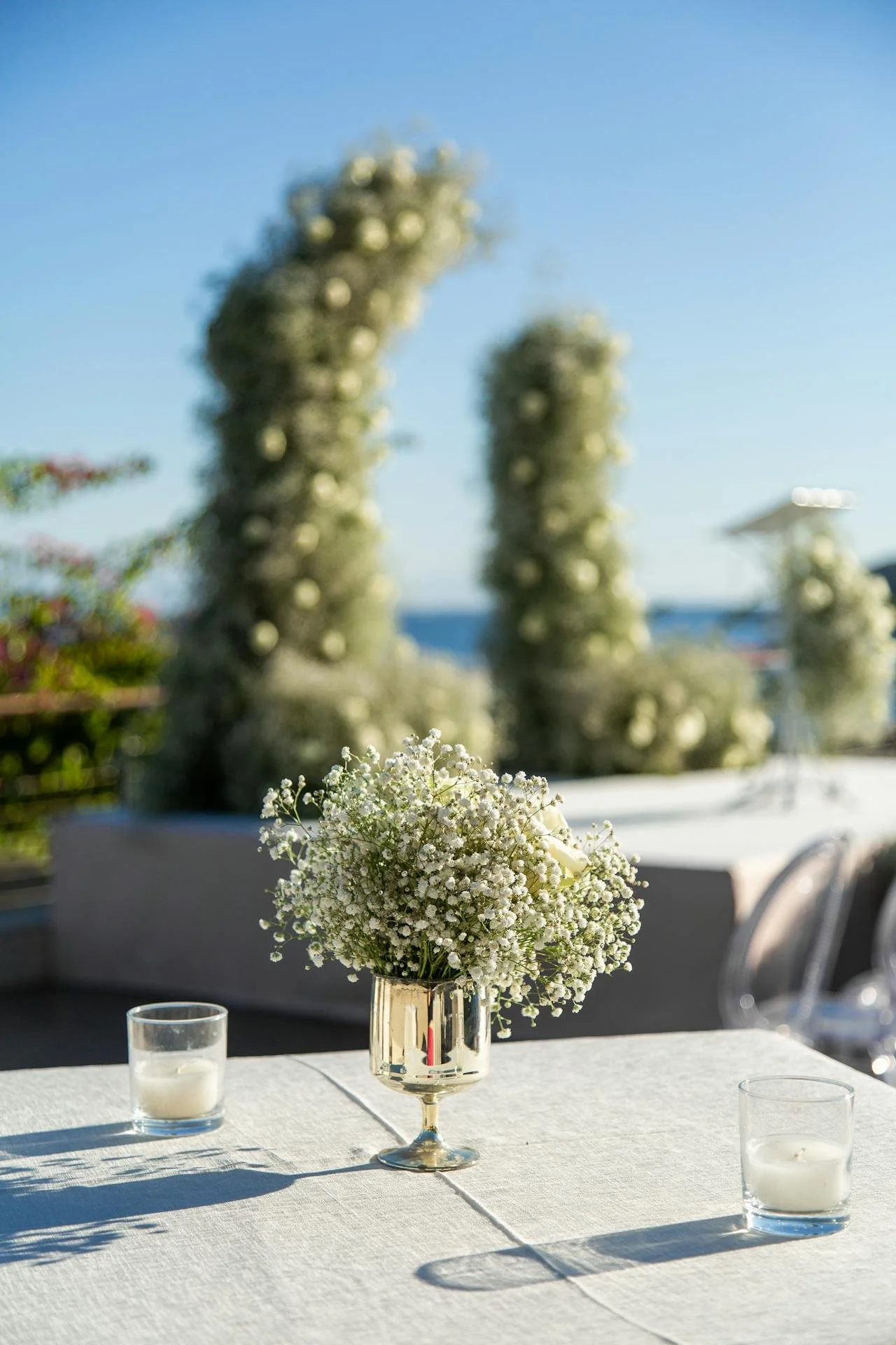 A table with a white tablecloth features a bouquet of white flowers in a reflective silver vase and two glass votive candle holders with white candles. The background shows a bright outdoor scene with blurred trees and the ocean under a clear blue sk