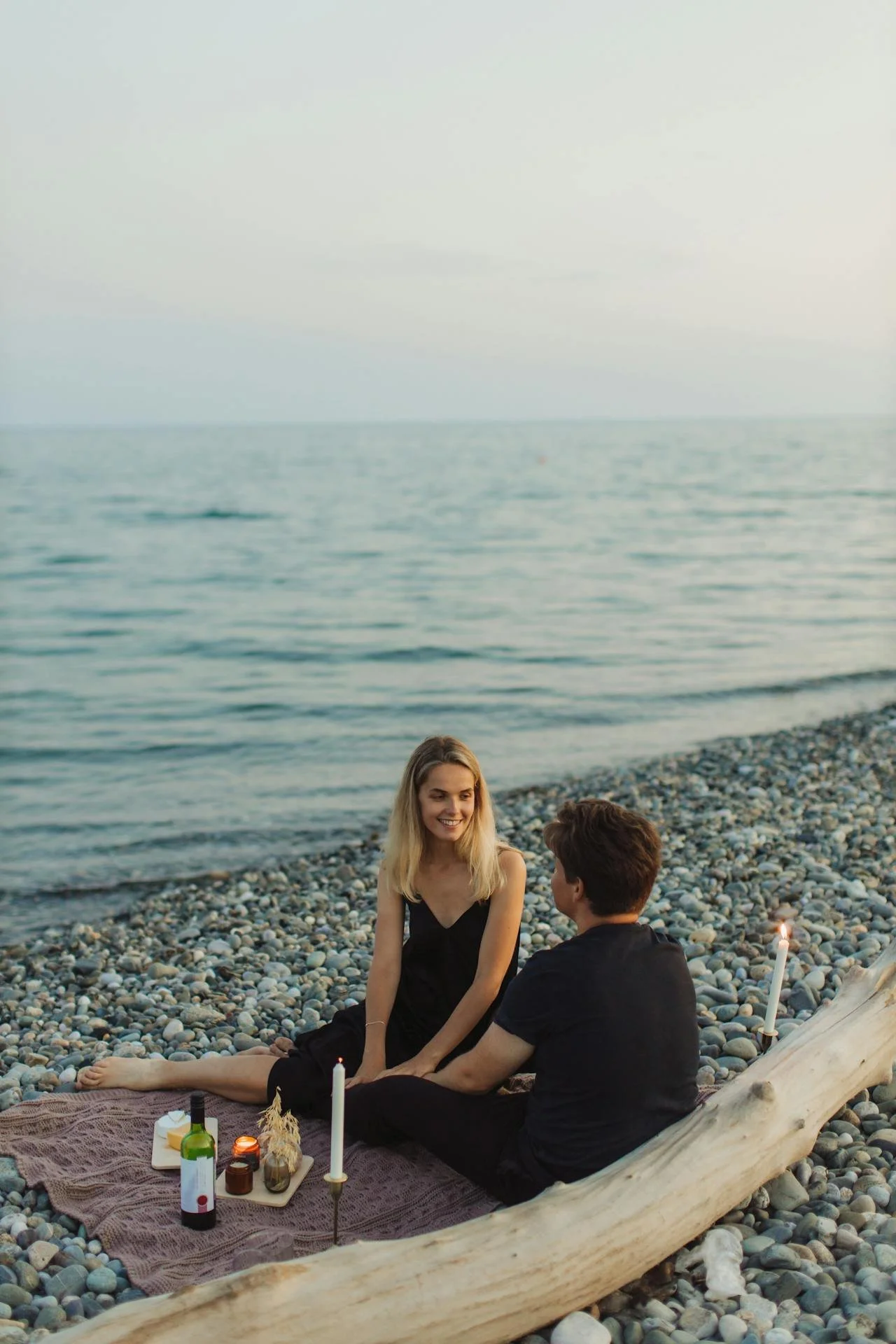 A couple sitting on a pebble beach during sunset with candles and a bottle of wine, engaging in conversation.
