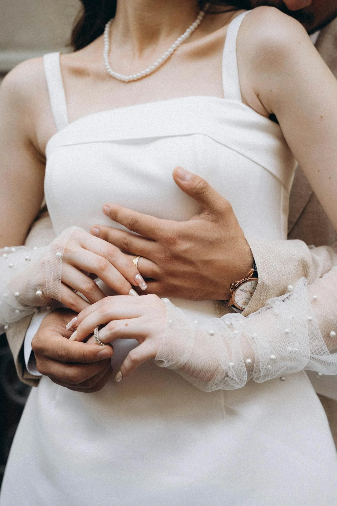 Close-up of a bride and groom holding hands, with the bride wearing a pearl necklace, a white wedding dress with sheer sleeves must decorated with pearls, and the groom wearing a watch and a gold ring.