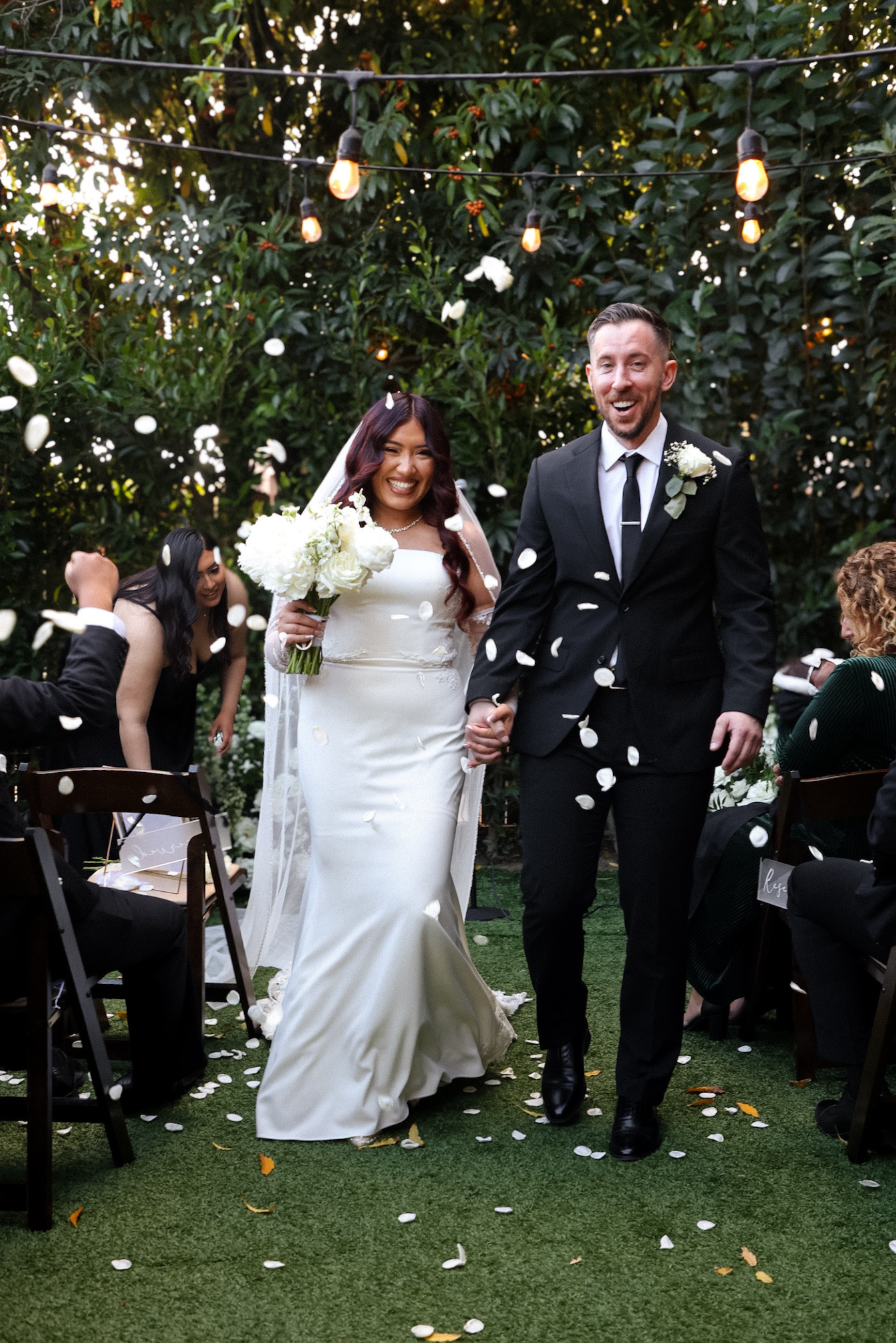 A newlywed couple walking hand in hand down an outdoor aisle, smiling, with guests throwing white flower petals around them during a wedding celebration.