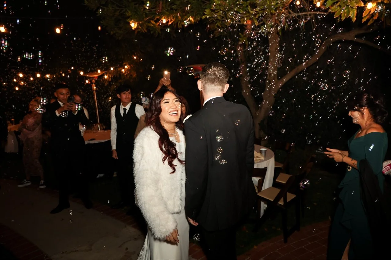 A wedding reception outdoors at night with fairy lights, a bride in a white outfit and a bridegroom in a dark suit smiling at each other, with guests taking photos and holding candles in the background.