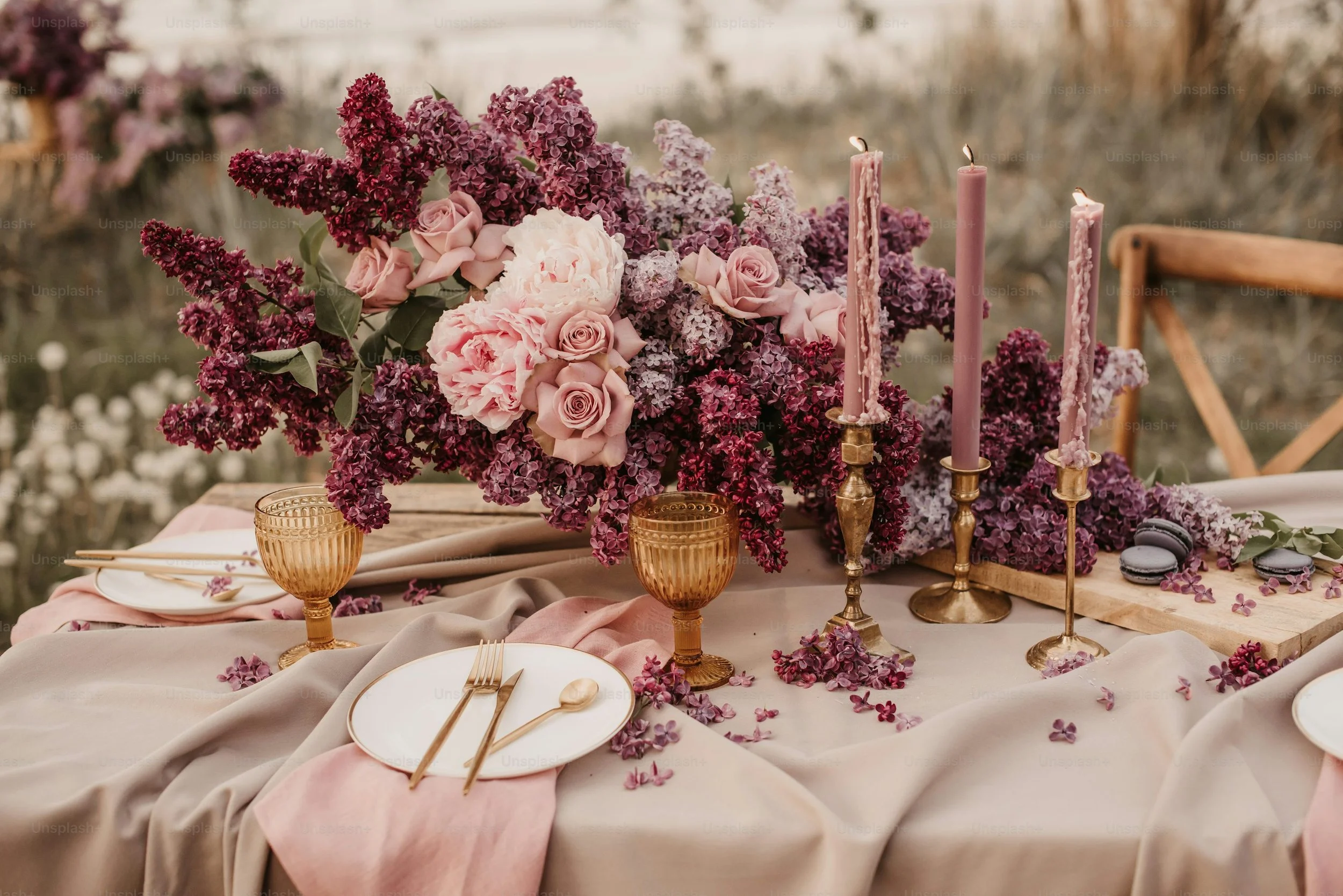 Elegant table setting with pink and purple floral arrangement, gold candle holders with pink candles, gold-rimmed plates, gold cutlery, copper-colored glasses, and macarons, set outdoors.