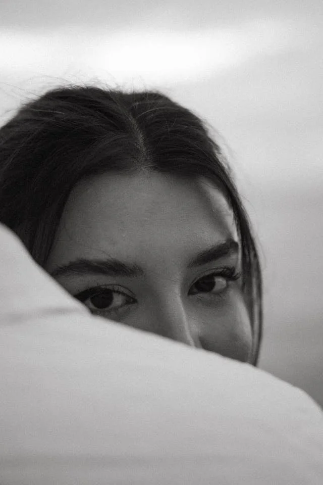 Black-and-white photo of a person's face partially hidden behind a pillow, showing only the eyes and part of the forehead.