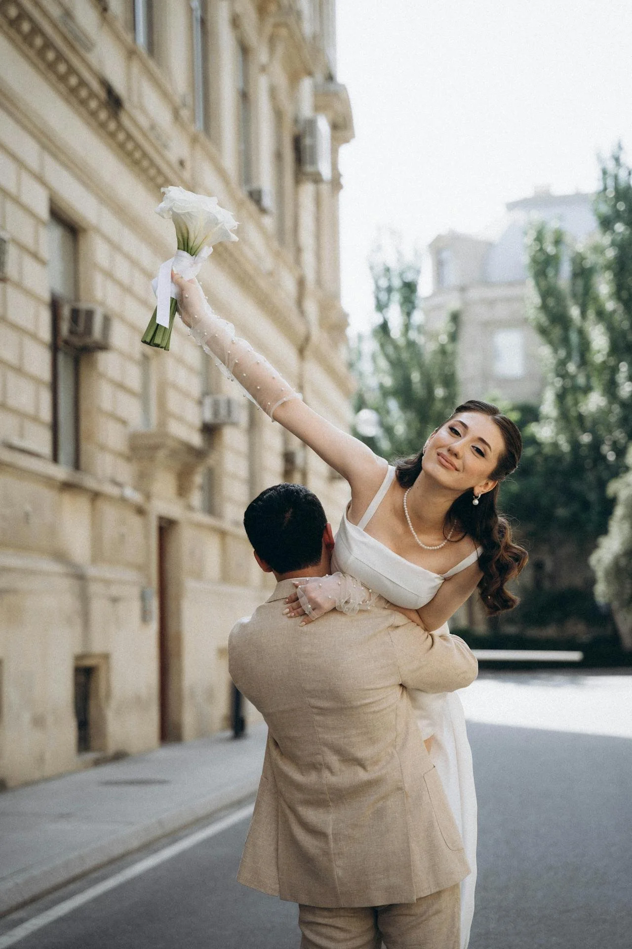 A couple dressed in wedding attire, with the woman holding a white flower bouquet, standing on a city street with historic buildings and greenery in the background.