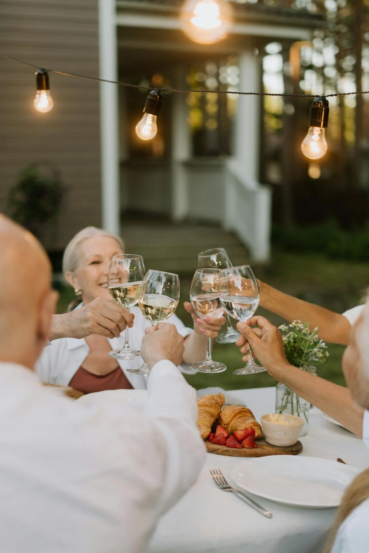 A group of seniors raising wine glasses in a toast at an outdoor dinner party under string lights, with a table of food including croissants and strawberries.