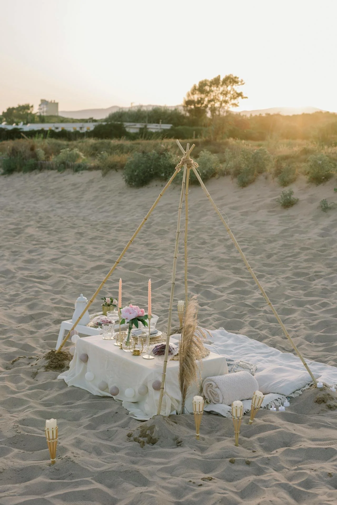 Beach picnic setup with a low table covered with a white cloth, decorated with candles, flowers, and blankets, arranged under bamboo poles, with tiki torches and candles around on sandy ground at sunset.