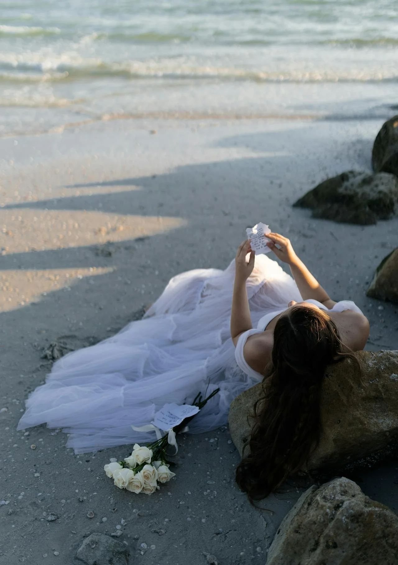 A woman in a white wedding dress lying on the sand near rocks at the beach, holding a crumpled note or letter, with a bouquet of white roses nearby.