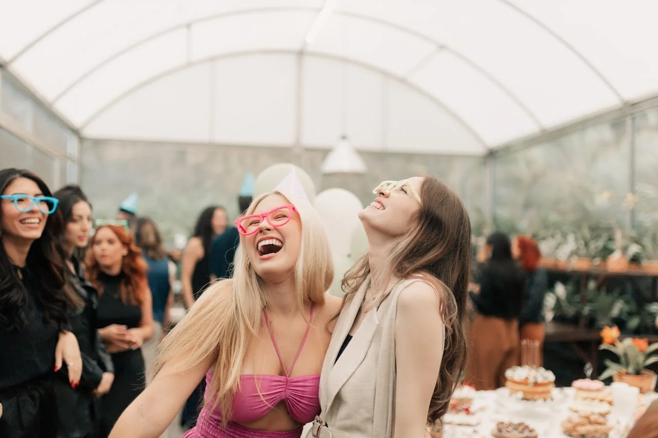 Group of women celebrating at a party, smiling and laughing, in a bright greenhouse with desserts on a table.