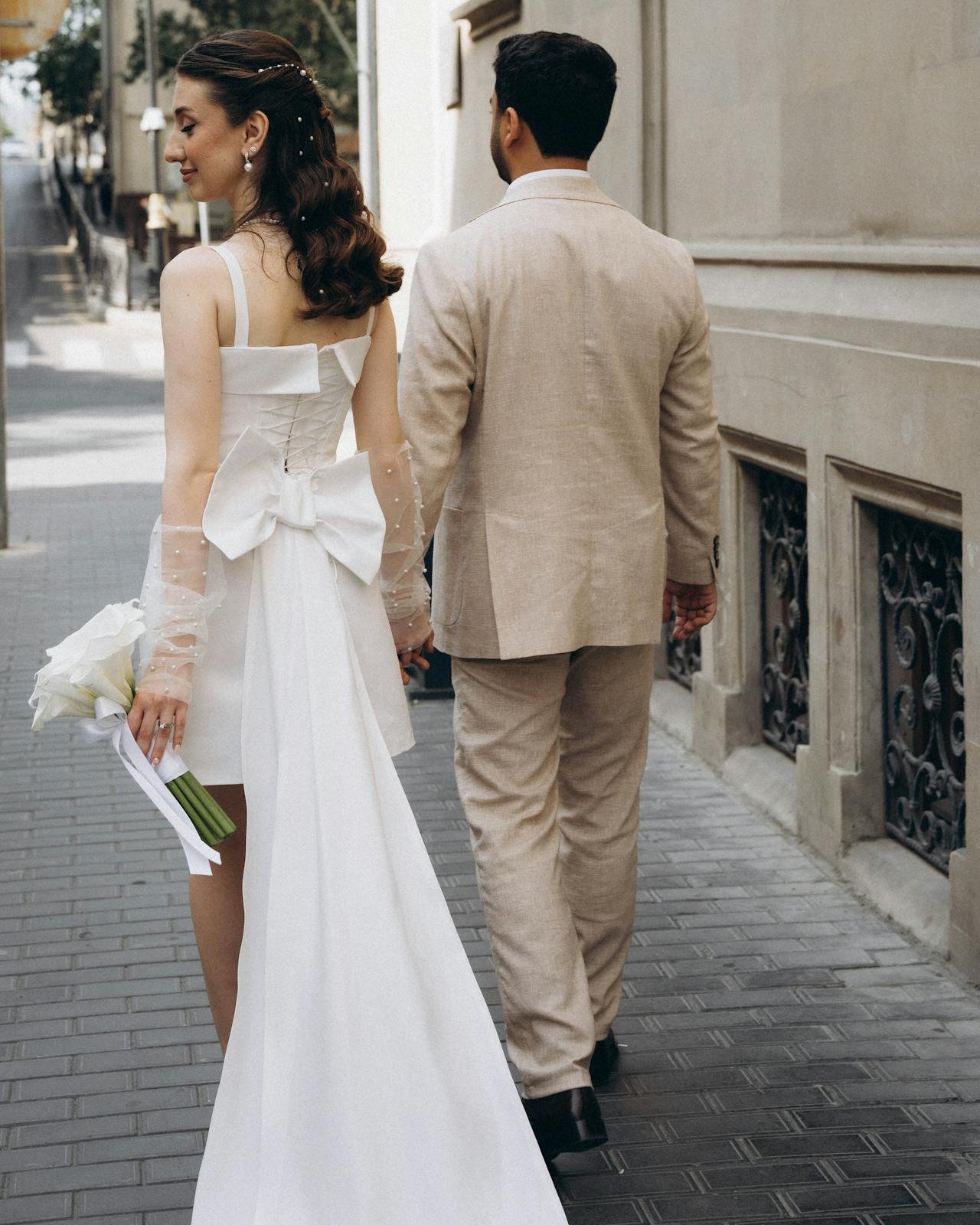A bride and groom walking hand in hand on a city sidewalk, the bride holding a bouquet of white calla lilies, she is wearing a white wedding gown with a large bow on the back, and the groom is in a beige suit.