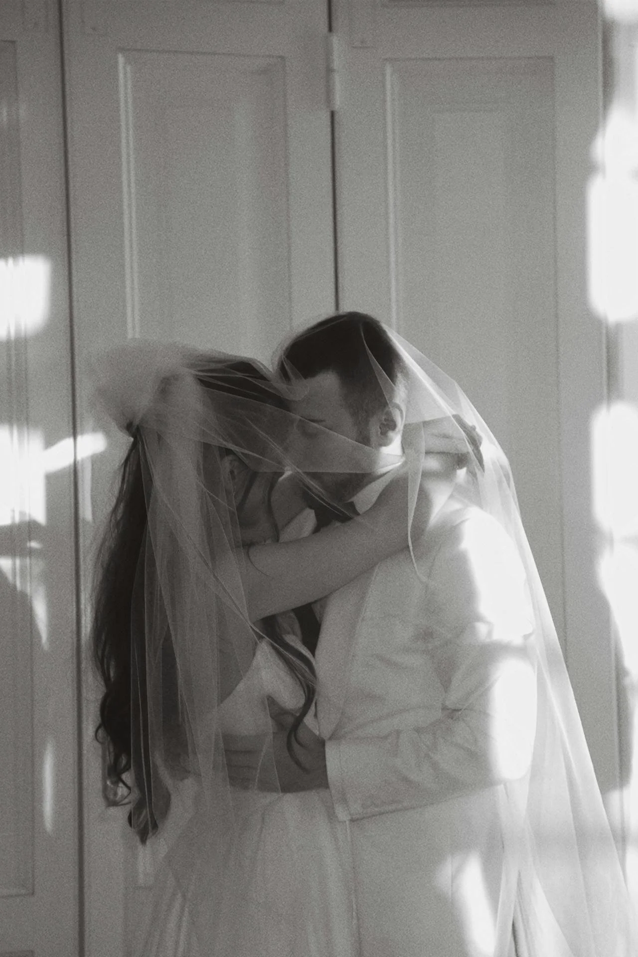 A black and white photo of a bride and groom embracing. The bride wears a veil and the groom wears a suit. They are close together with their faces touching, standing against a plain background with shadows cast on the wall.