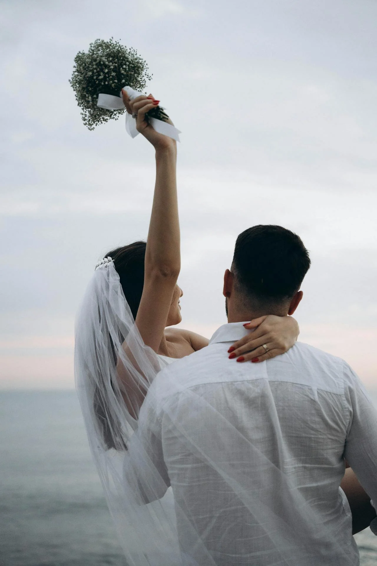 A bride holding a bouquet of flowers above her head while embracing her groom, overlooking the ocean during the evening.