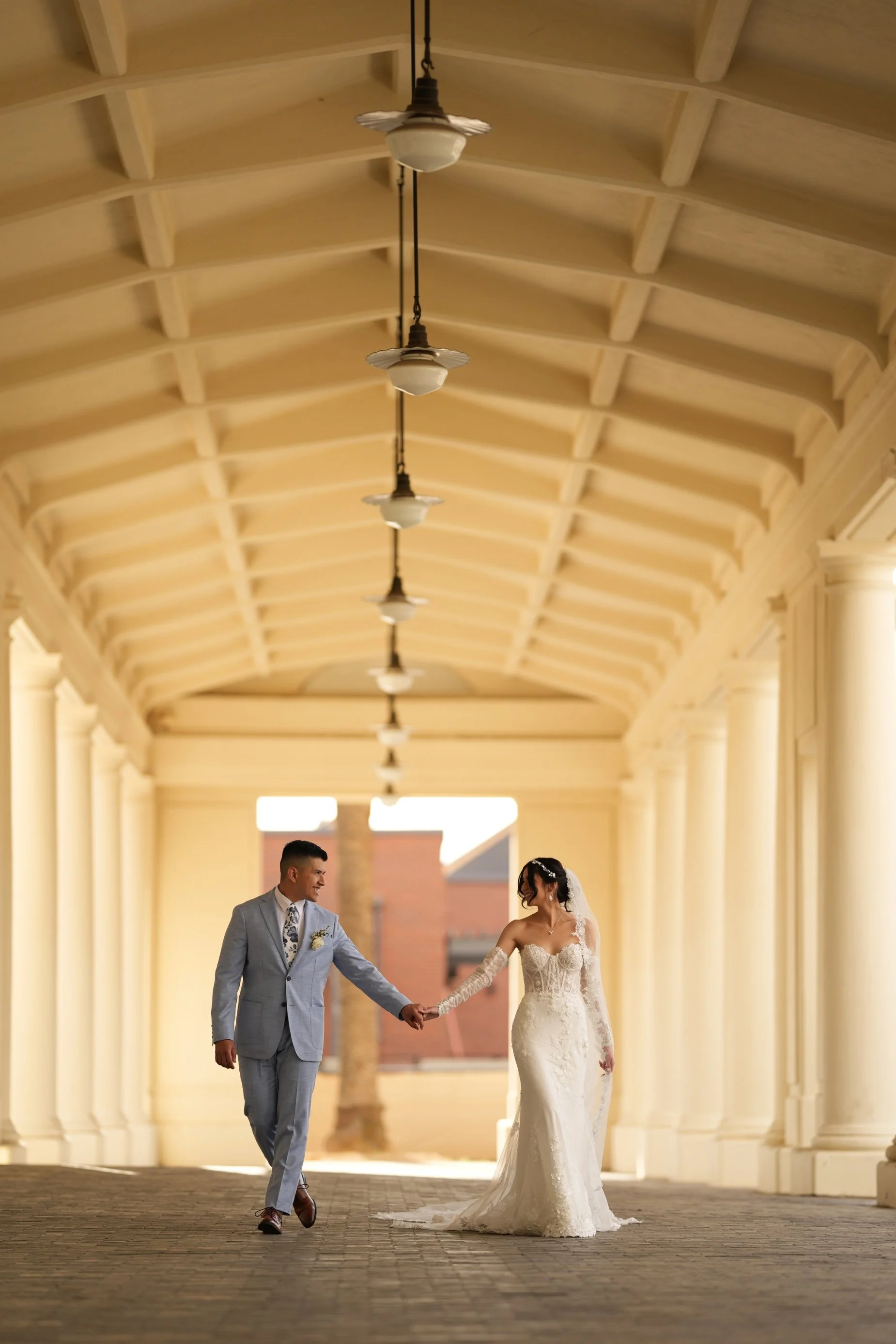 A bride and groom holding hands and walking in an elegant, light-colored, covered walkway.
