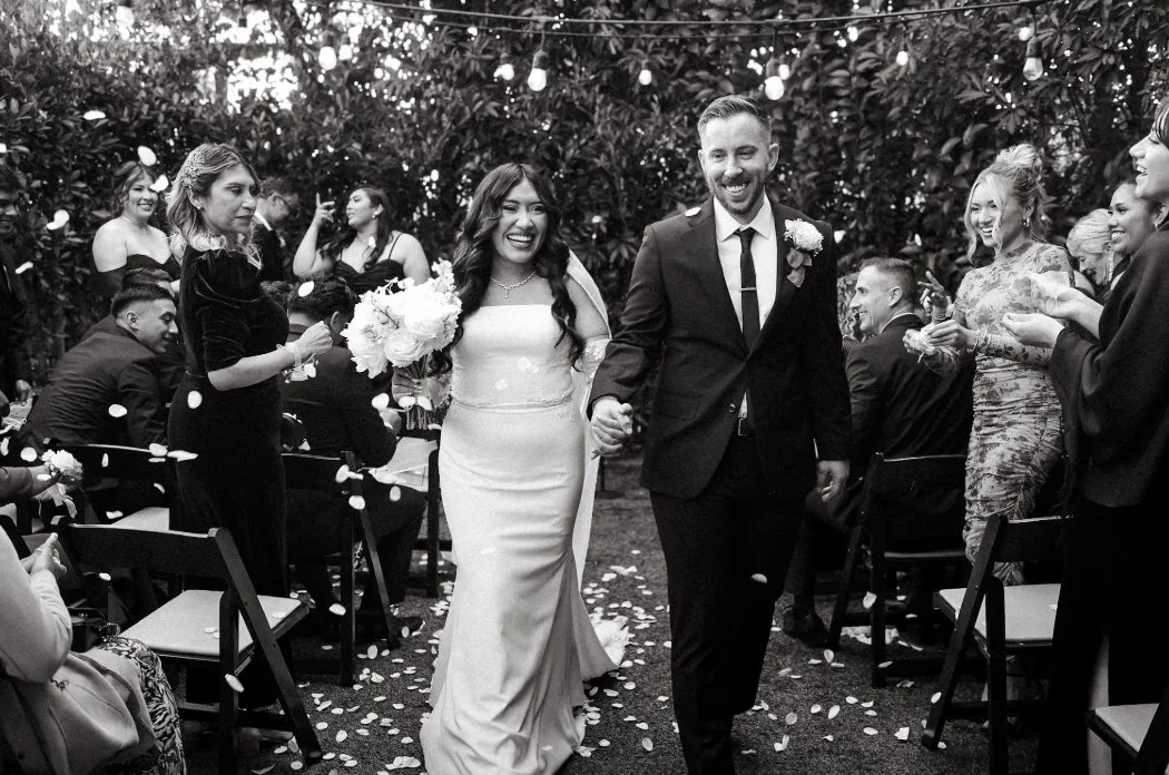 A black and white photograph of a wedding celebration, featuring a bride and groom walking hand in hand down the aisle, smiling and surrounded by seated and standing guests cheering and celebrating.
