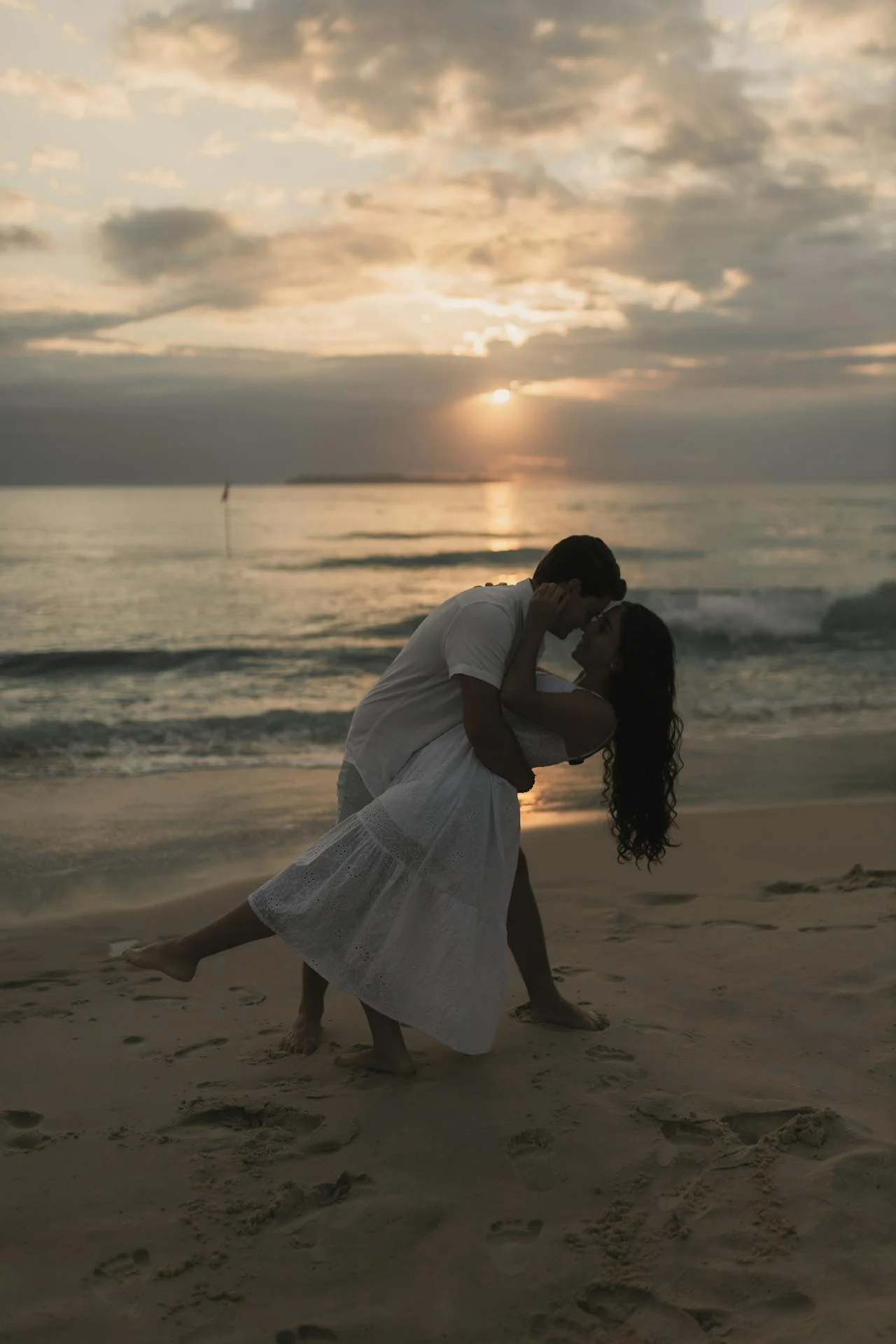 A couple embracing on a beach at sunset, with the man dipping the woman as they kiss, the ocean and cloudy sky in the background.