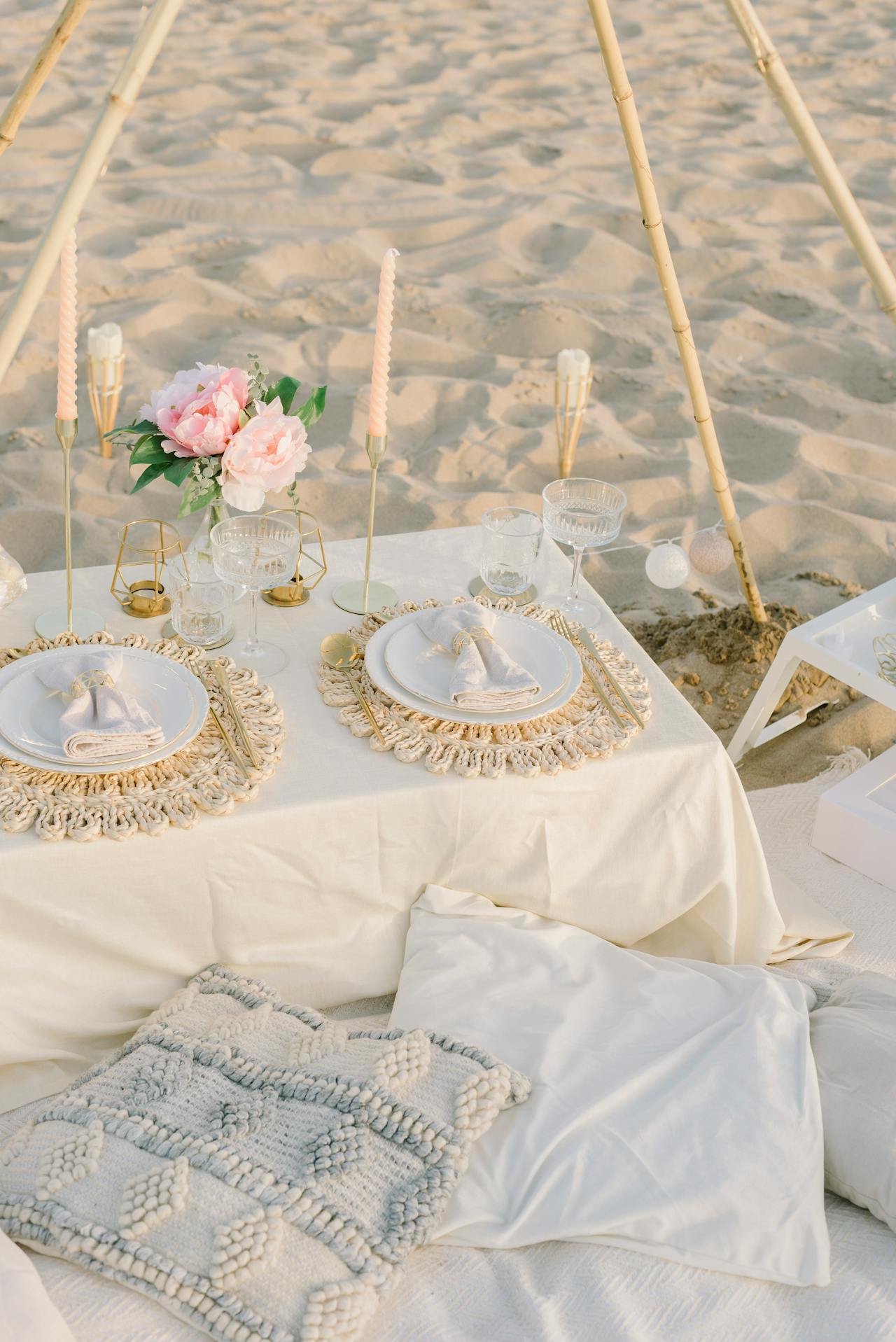 Beachside table setting with white tablecloth, pink floral centerpiece, candles, glassware, and cloth napkins, under a bamboo canopy.