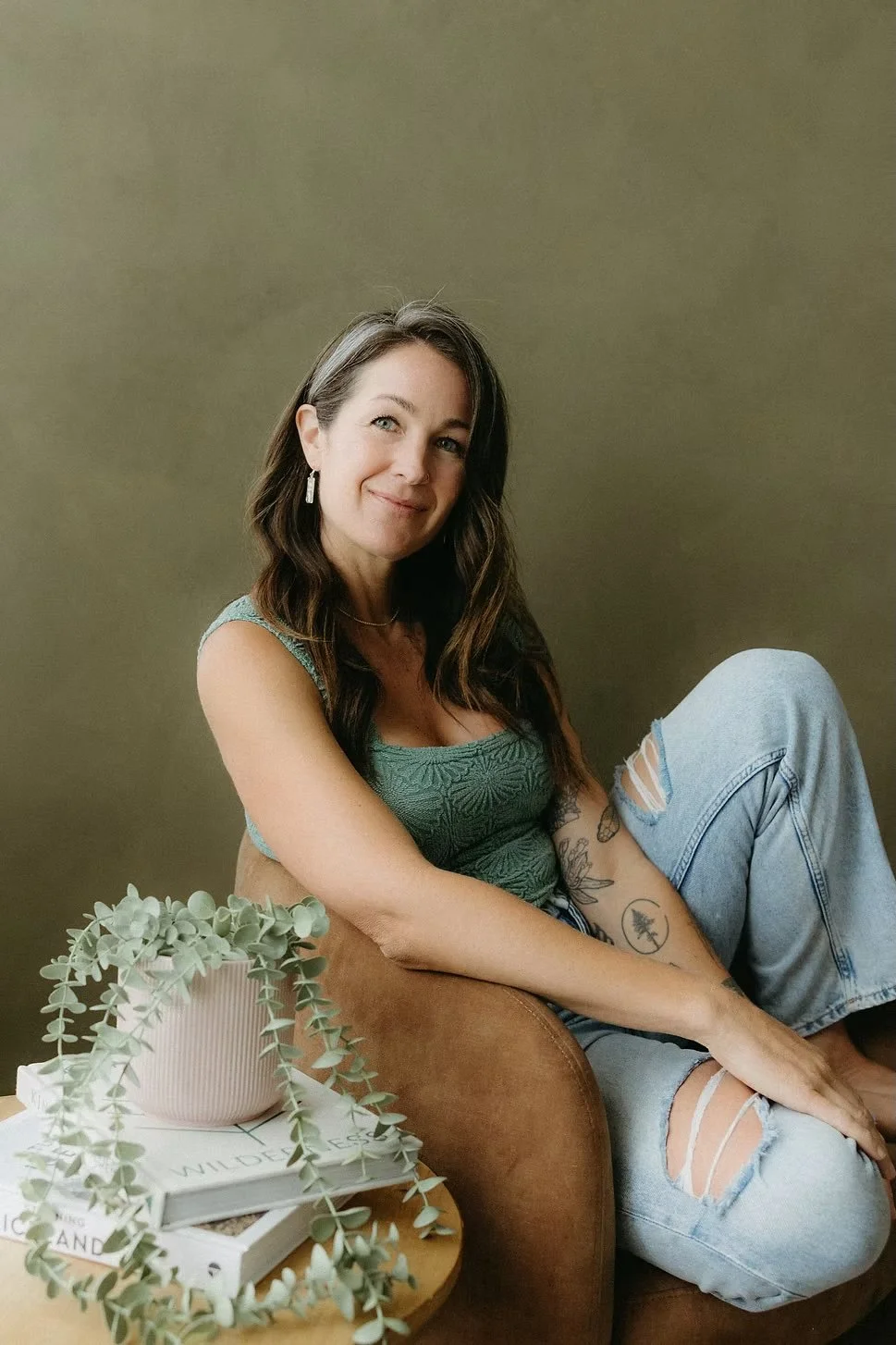 A woman with long dark hair and tattoos sitting on a brown armchair next to a small round table with a pink planter and a couple of books, wearing a green sleeveless top and ripped jeans, smiling at the camera against a plain wall background.