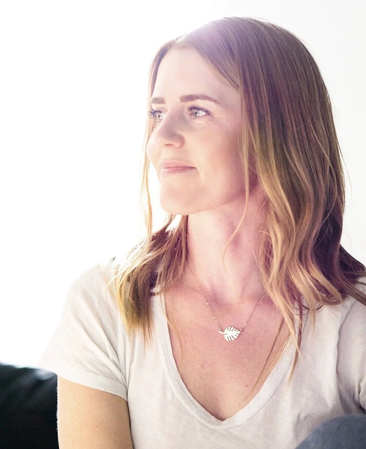 A woman with auburn hair and a light complexion looking to her left with a soft smile, wearing a white t-shirt and a necklace with a leaf-shaped pendant.