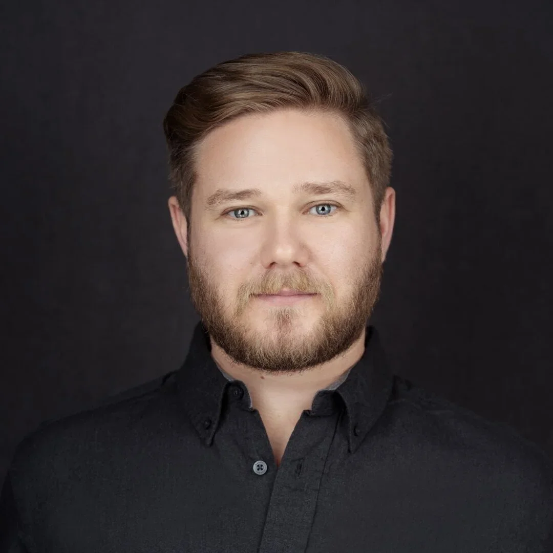 A young man with blonde hair and a beard, wearing a black shirt, against a dark background.