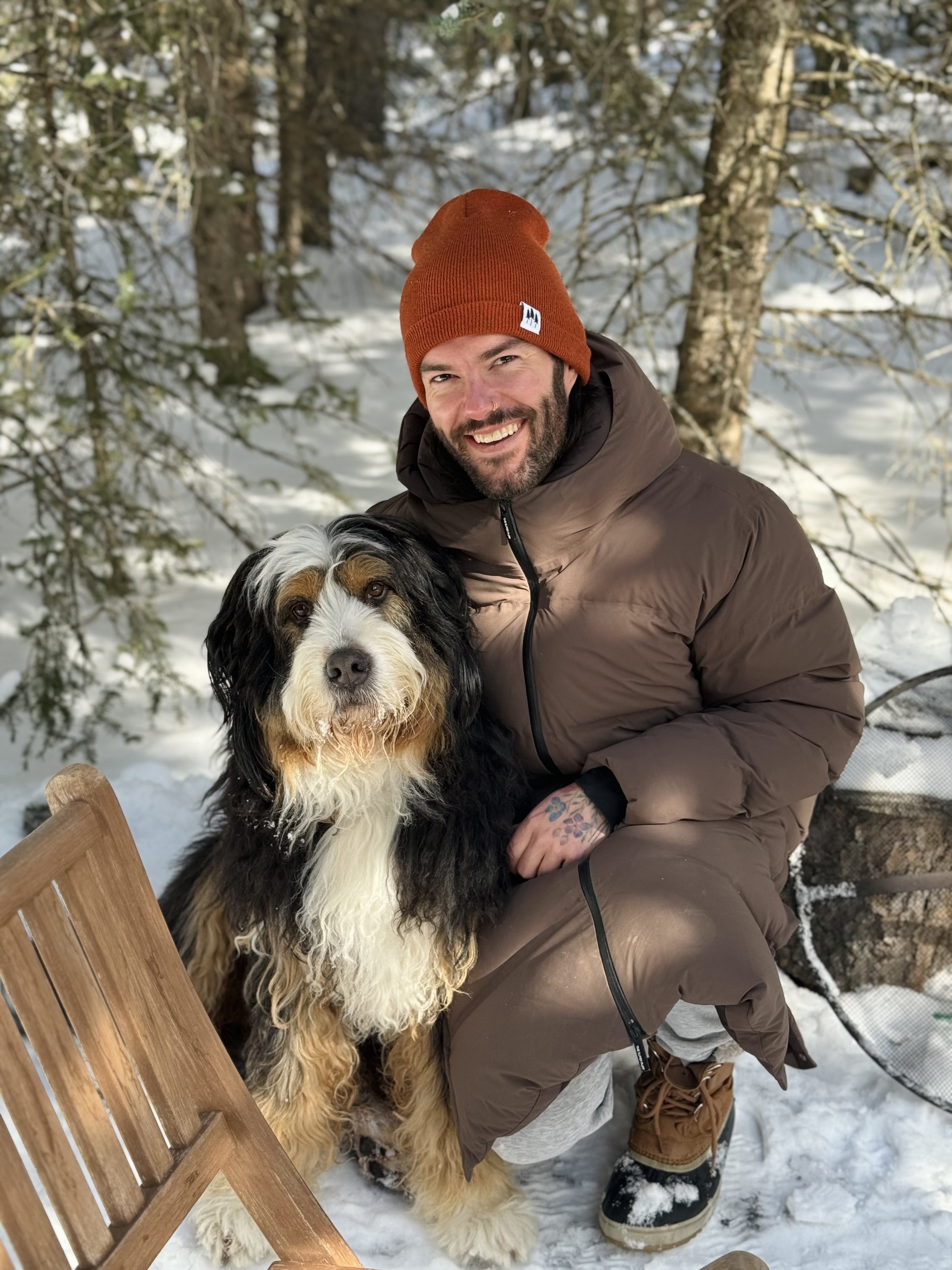 A smiling man with a beard, wearing a brown winter coat, gray pants, and a red beanie, kneels beside a large, black, white, and tan Bernese Mountain dog in a snowy outdoor setting with trees.