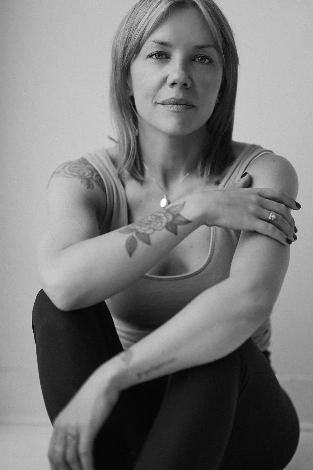 Black and white portrait of a woman with shoulder-length hair, tattoos on her arms, wearing a sleeveless top, sitting with her arms crossed over her knees, looking directly at the camera.