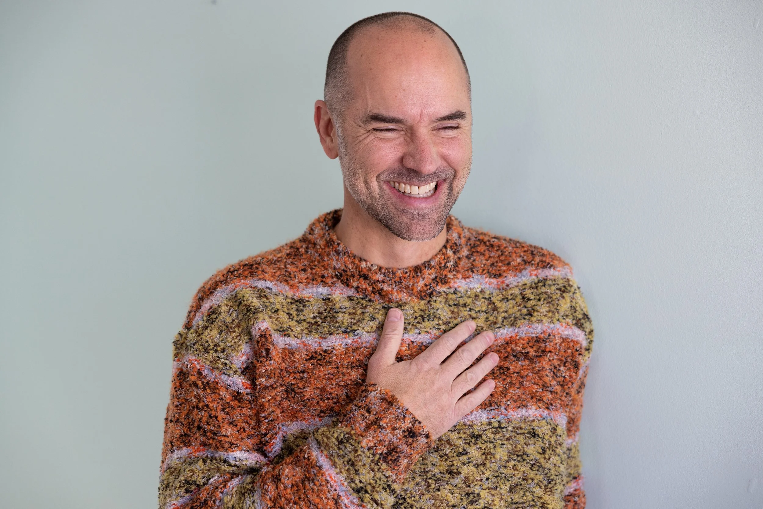 A middle-aged man with a bald head and gray beard, smiling with his eyes closed, touching his chest with his right hand, wearing a colorful, textured sweater, standing against a plain, light-colored wall.