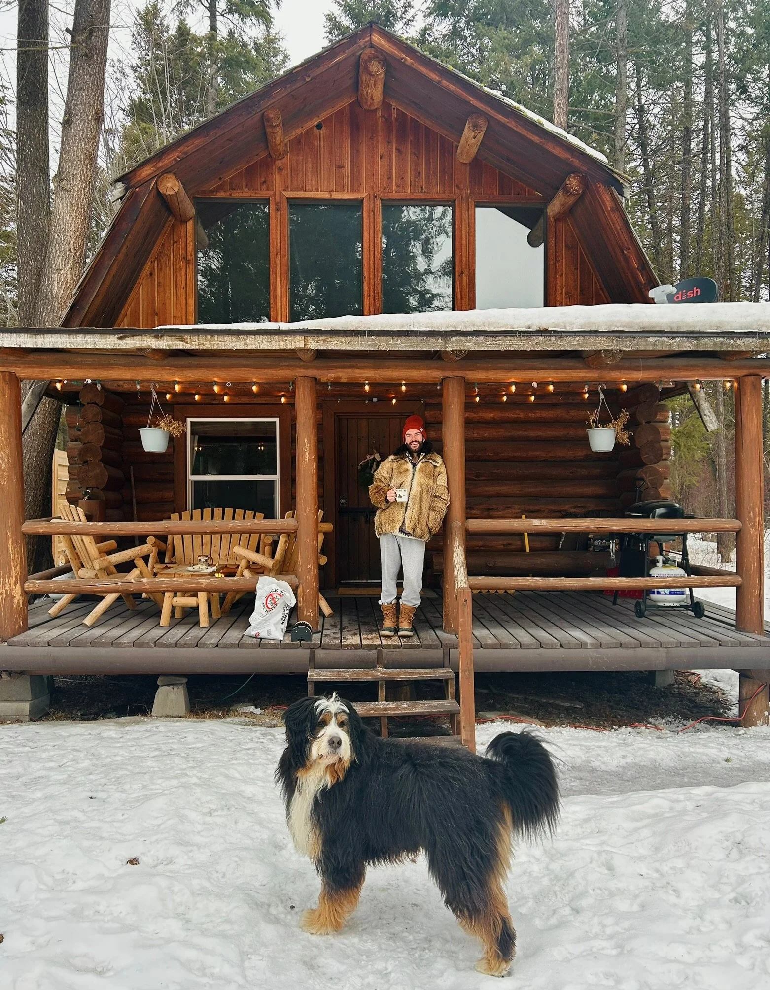 A person standing on the porch of a log cabin holding a mug, with a dog standing on snowy ground in front.