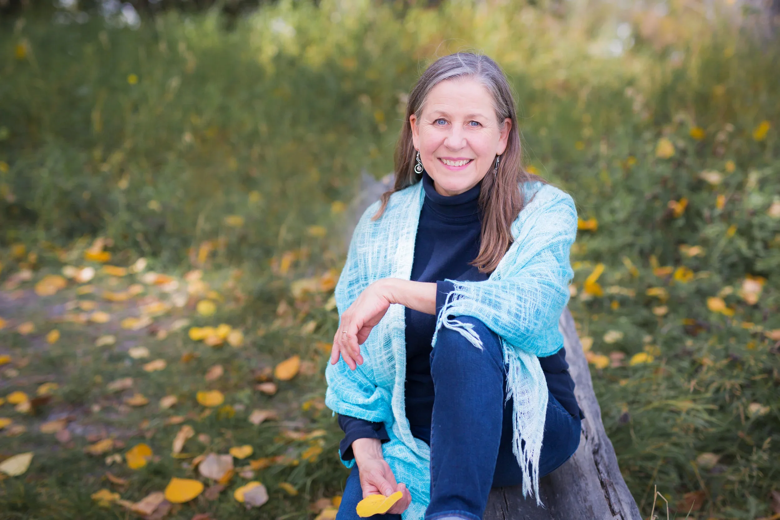 A smiling woman sitting outdoors on a fallen tree trunk in a park during autumn, wearing a black turtleneck, blue jeans, and a light blue shawl, holding a yellow leaf in her right hand.
