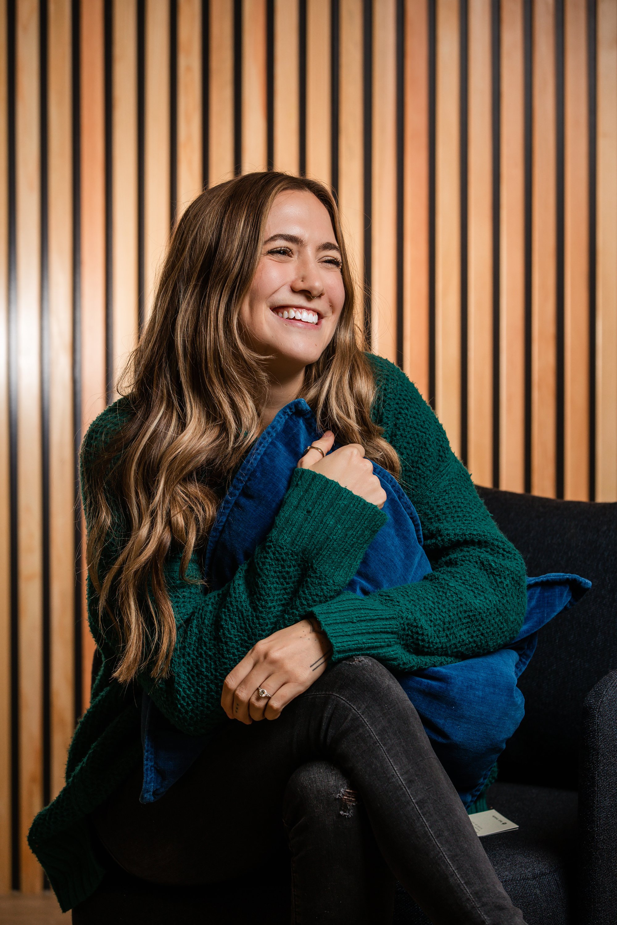 A young woman with long wavy brown hair smiling and hugging a blue pillow, sitting on a black couch in front of a wooden slat wall.