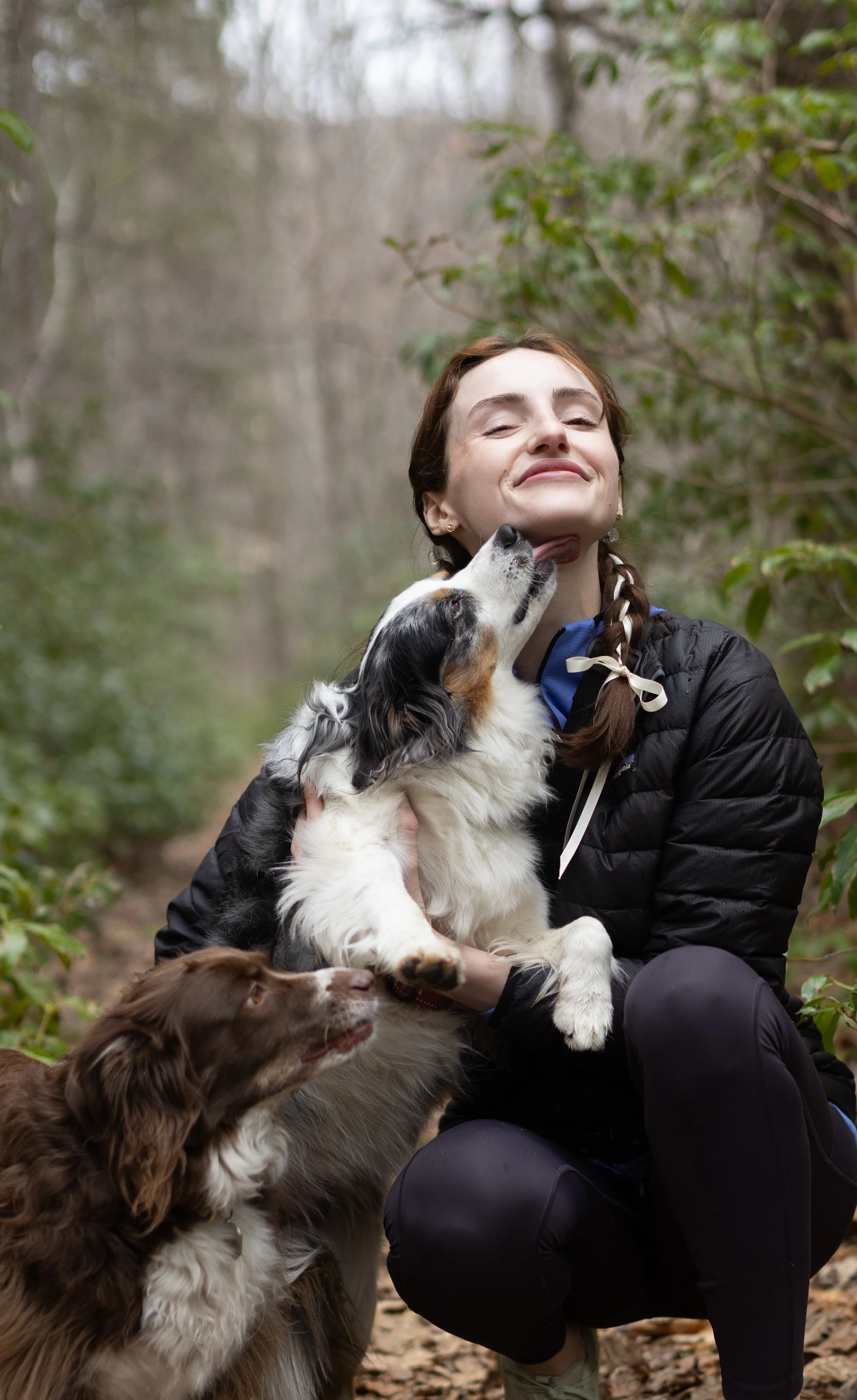 A woman smiling while holding an Australian Shepherd puppy, with another dog next to her, in a wooded outdoor setting.