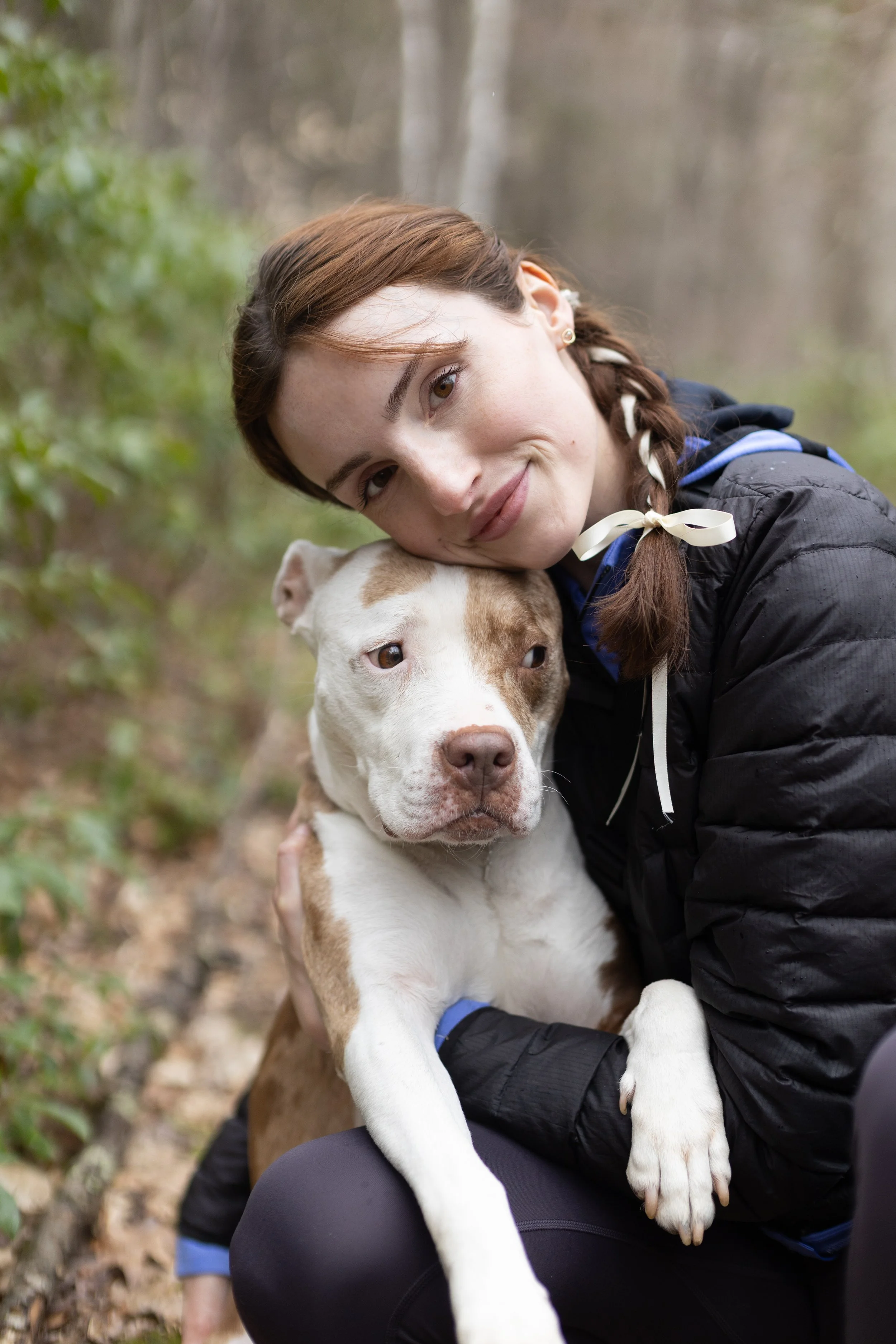 A woman with red hair in a braid, wearing a black jacket, hugging a brown and white dog in a wooded outdoor area.