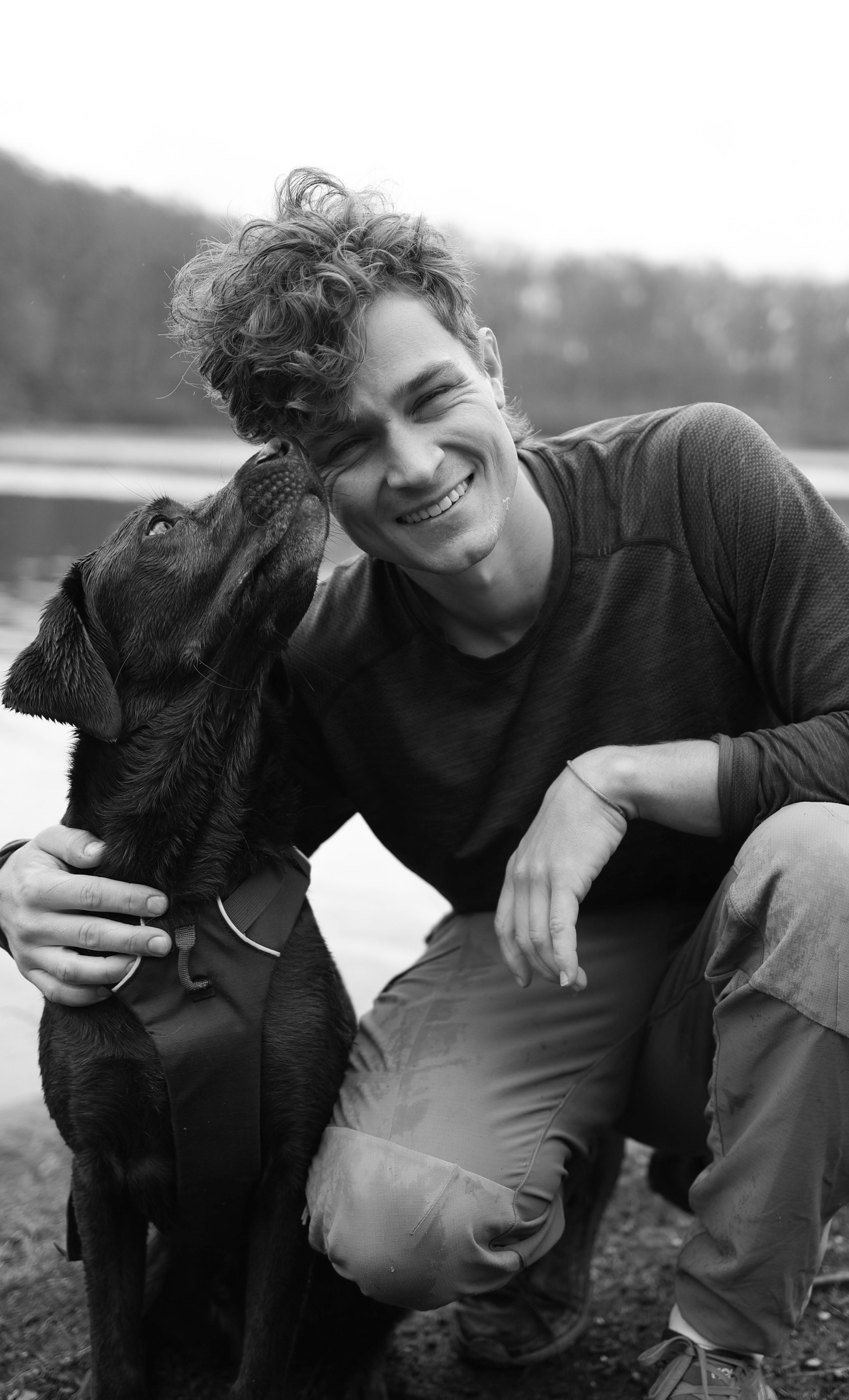 A man with curly hair smiling and petting a Labrador retriever outdoors near a body of water.