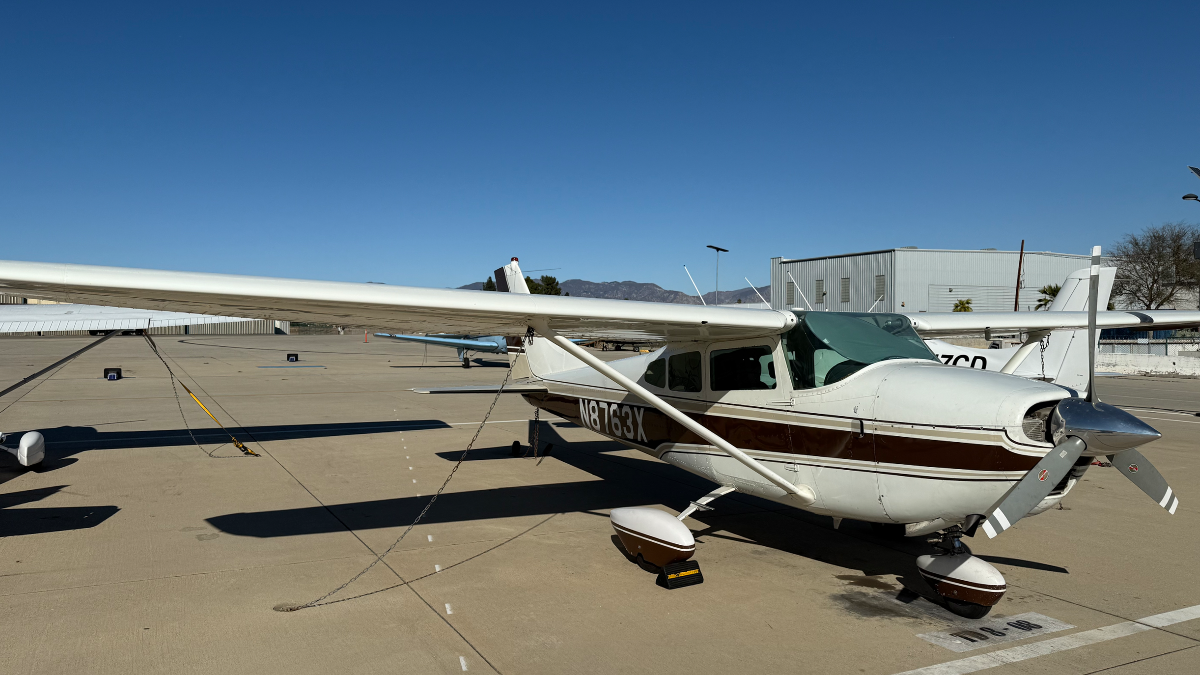 Small white propeller airplane parked on tarmac at airport, tied down with chains, under clear blue sky.