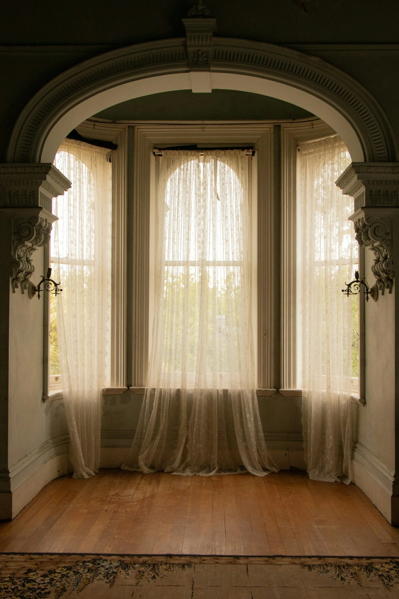 Sunlit room with large arched window featuring sheer lace curtains, ornate architectural details, and wooden floor.