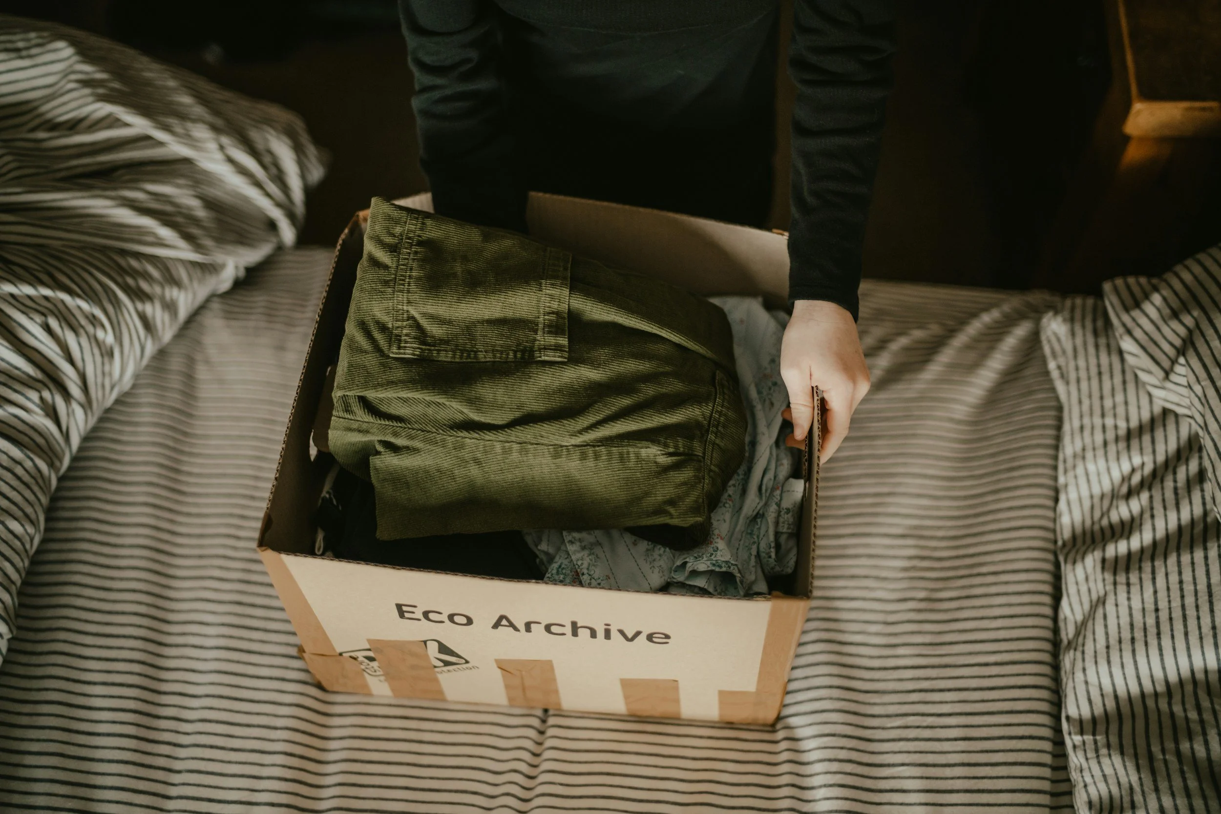 Person packing clothes into a cardboard box labeled 'Eco Archive' on a bed with striped bedding.