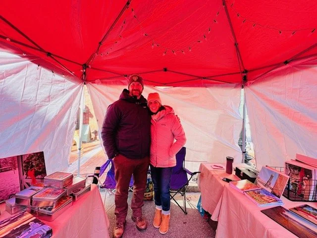 A man and woman standing inside a red and white tent at a market or fair, with tables displaying baked goods and books, and decorations hanging from the ceiling.