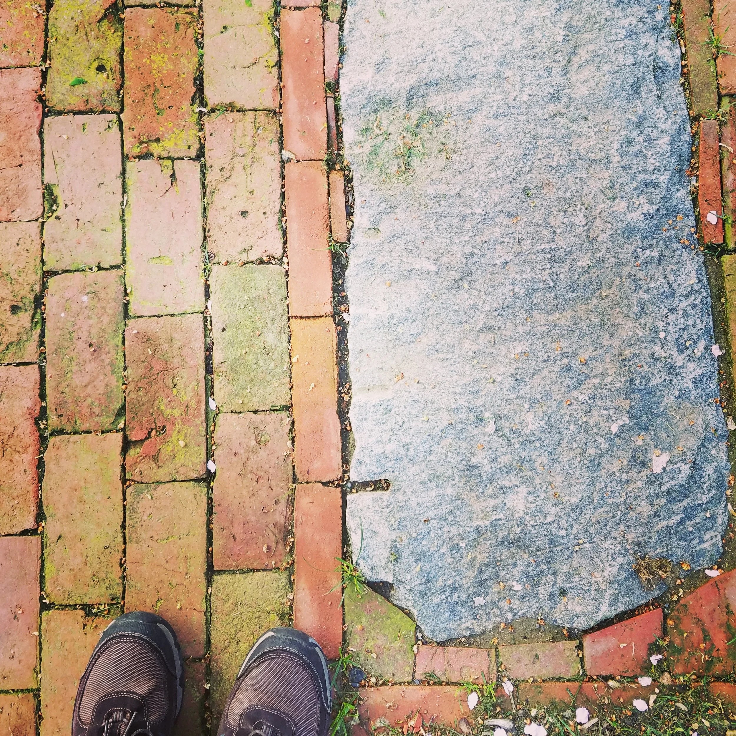 Close-up of person's feet in black shoes standing on a stone pathway with red and green moss-covered bricks, next to a large worn gray stone slab.