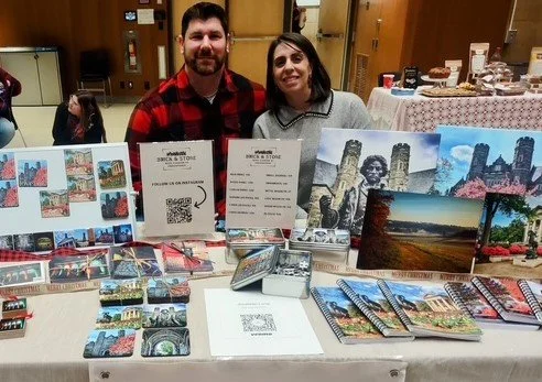 Two people standing behind a table displaying travel brochures and photos, with various brochures, postcards, and imagery of scenic landscapes and cityscapes.