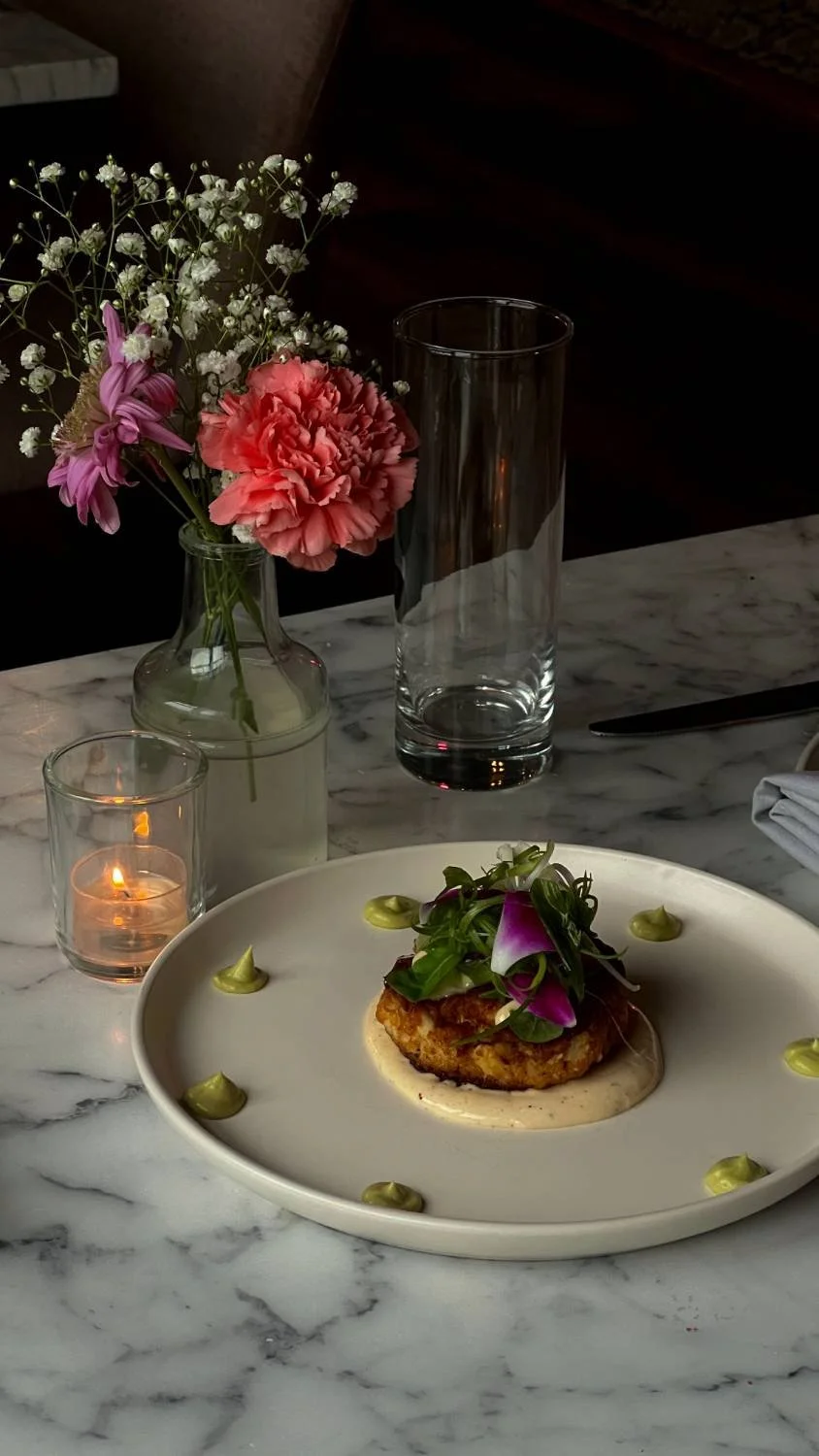 A dish with a fried patty topped with greens and edible flowers, served on a white plate with green sauce decorations around the edge. In the background, a floral arrangement with pink and purple flowers, a lit candle, and an empty glass vase are on 