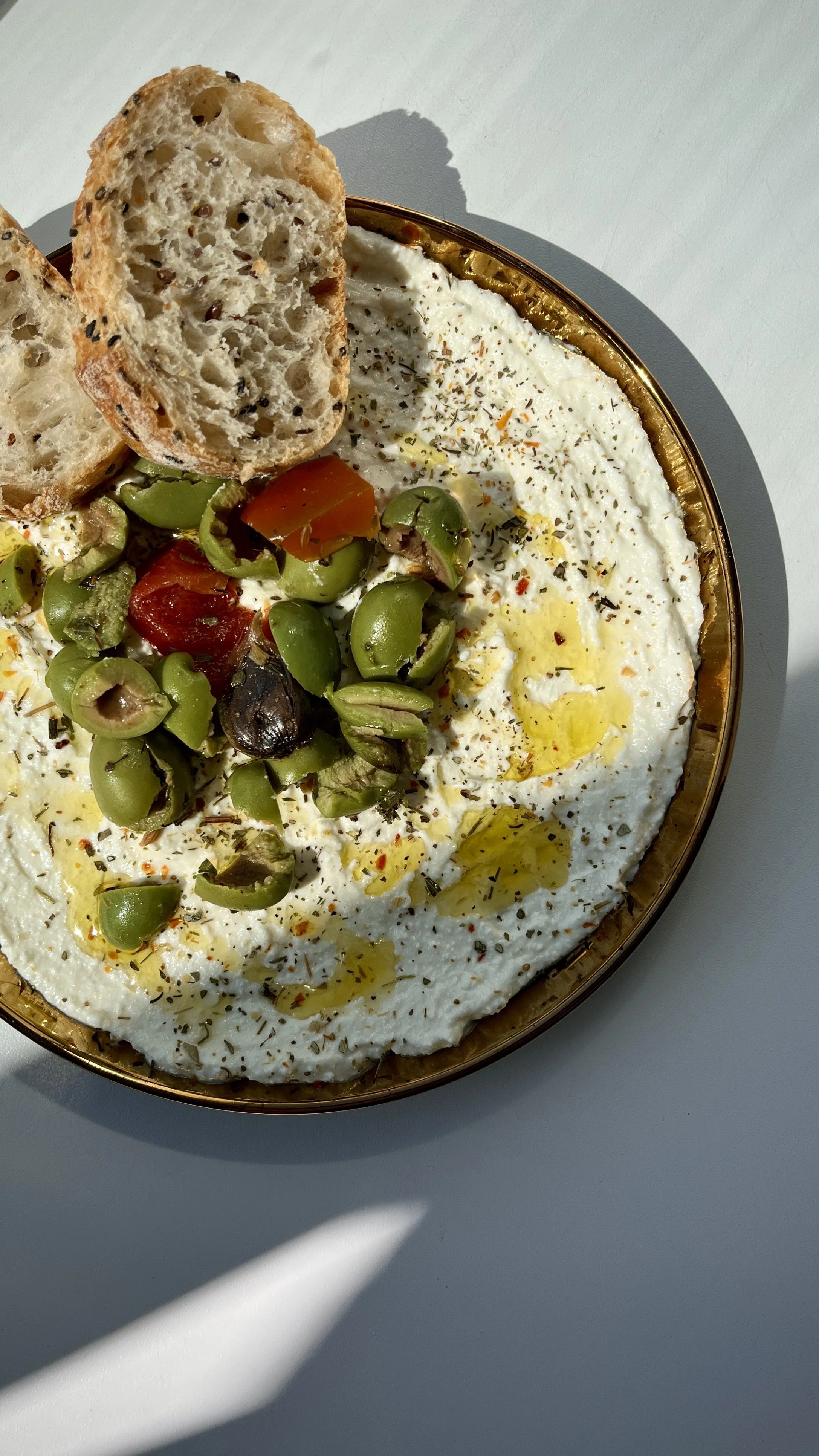 Gourmet Gals Catering a plate of hummus garnished with herbs, olive oil, and spices, served with green olives and chopped vegetables, with toasted bread slices.