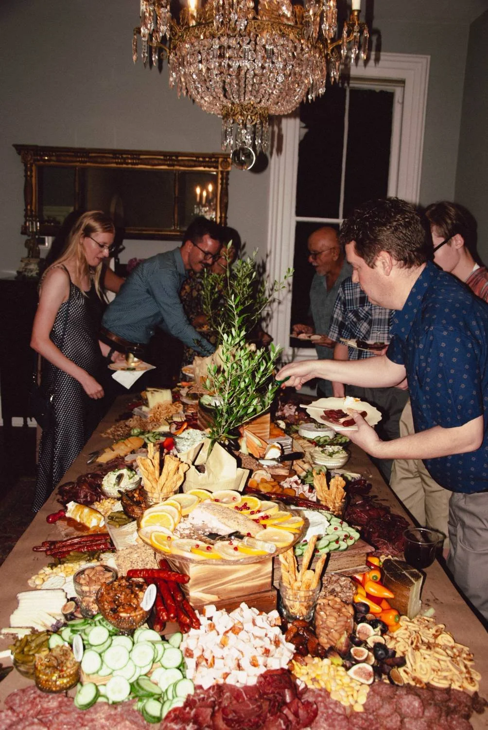 People serving themselves food from a buffet table in a dining room with a large chandelier and a mirror.