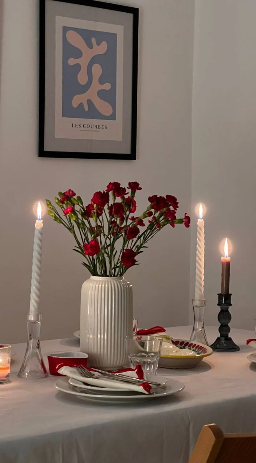 A dining table decorated for a meal with a large white vase holding red flowers as the centerpiece, surrounded by lit candles, plates, glasses, and utensils, with framed artwork titled "Les Courbes" on the wall behind.