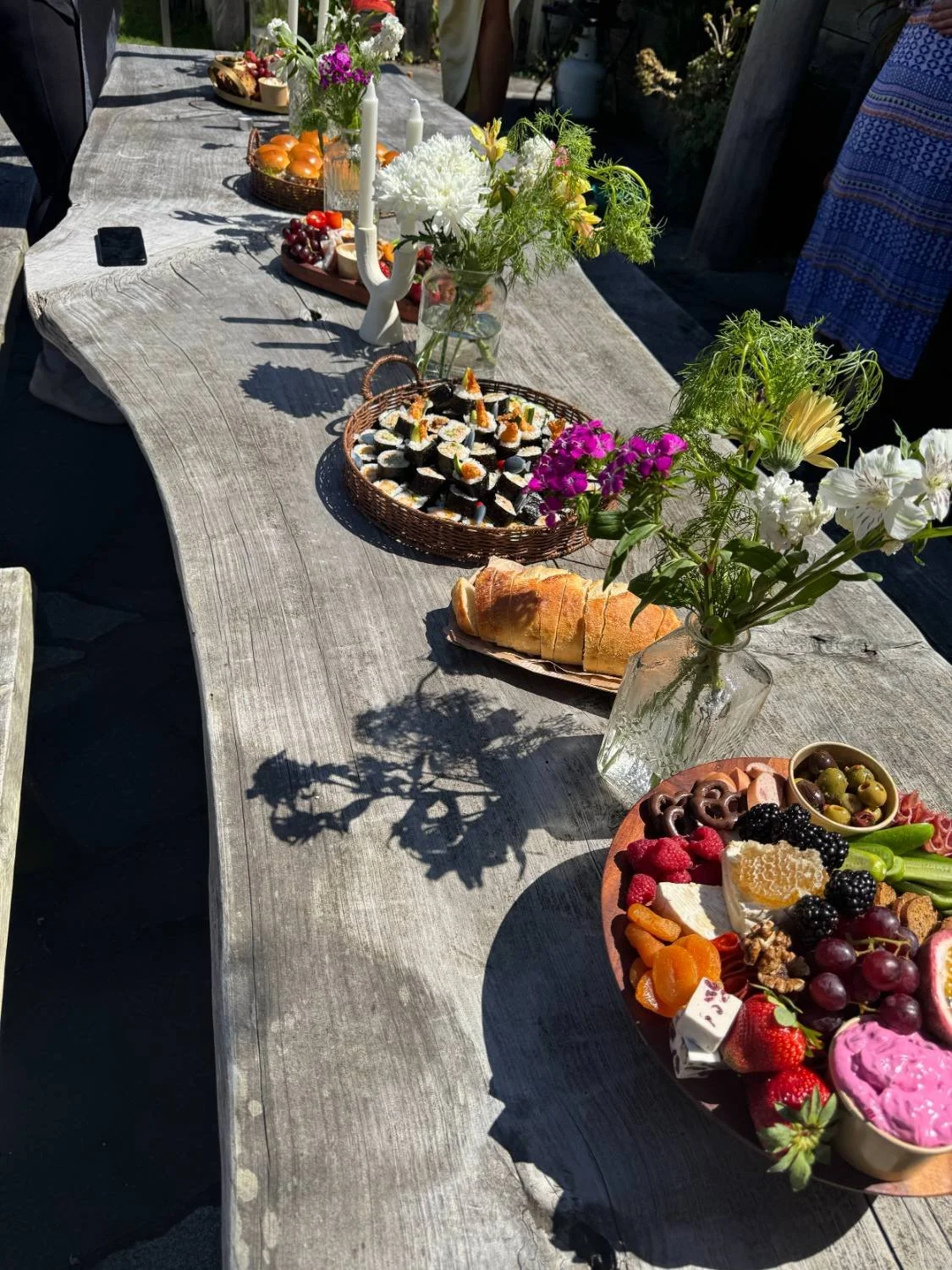 An outdoor table with a rustic wooden surface set for a gathering, decorated with vases of colorful flowers, plates of sushi, bread, and an array of fruits, nuts, and desserts.