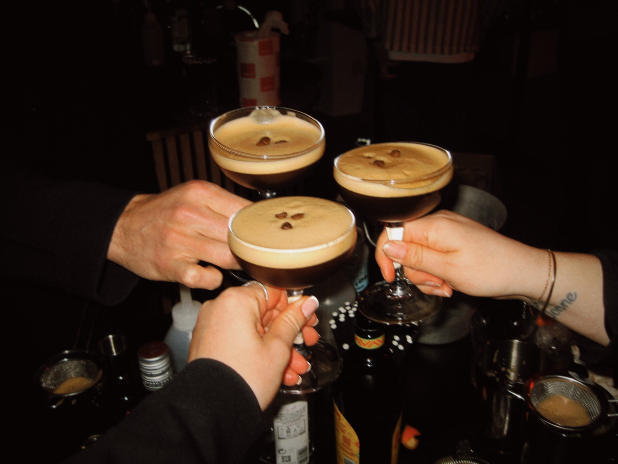 Three people clinking glasses filled with espresso martinis in a dark setting.