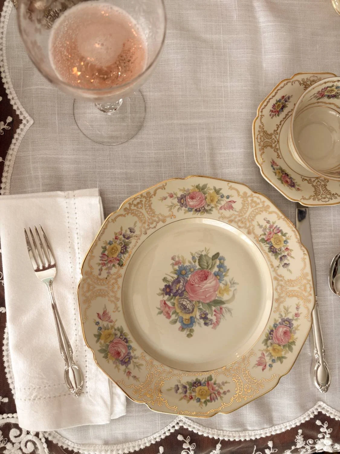 A formal dining table setting with a floral china plate, a matching cup and saucer, a water glass with rosé wine, a silver fork on a white napkin, and silver knife, on a white tablecloth with lace trim.