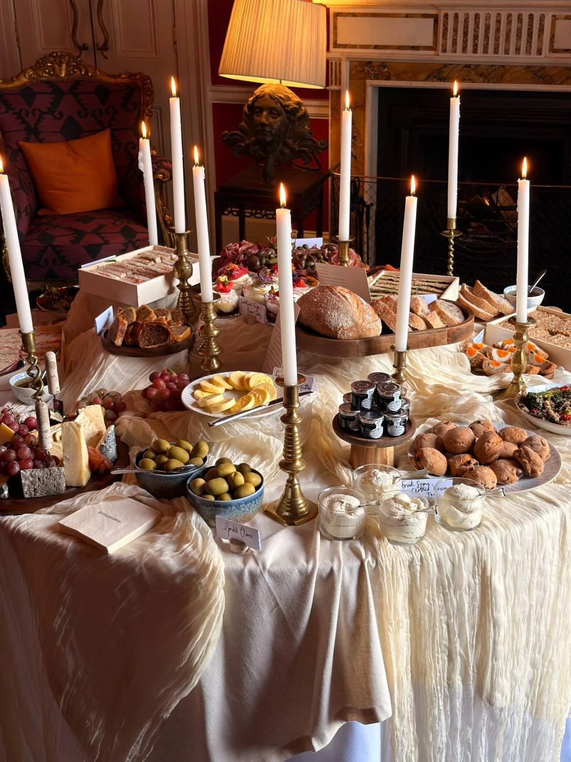 A formal dining table set with candles, bread, cheese, grapes, olives, and desserts for a festive occasion.